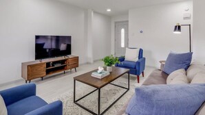 A modern living room with a flat-screen TV, a wooden entertainment unit, a coffee table, a blue sofa, and a beige sofa. - Best of Irving - Yard, Firepit, Free Parking (Irving)