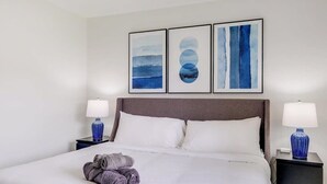 A neatly made bed with white linens, two pillows, and a stack of rolled towels on the foot of the bed. There are two bedside tables with lamps, and three framed artworks above the bed. - Best of Irving - Yard, Firepit, Free Parking (Irving)