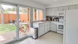 A modern kitchen with white cabinets, a stainless steel range hood, and a built-in oven. There is a sliding glass door leading to a patio area with outdoor furniture. - Best of Irving - Yard, Firepit, Free Parking (Irving)