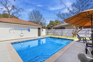 Ein rechteckiger Swimmingpool mit blauer Fliesenbordüre, umgeben von einer Betonplattform. Liegestühle und ein Tisch befinden sich unter einem Sonnensegel.