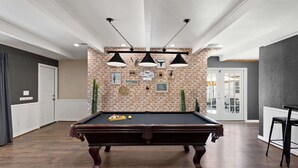 Een pooltafel in een ruimte met bakstenen muren, hanglampen en een deur die naar een andere zone leidt.