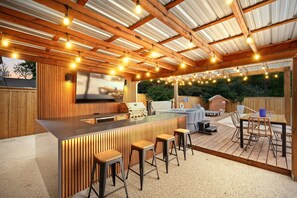 An outdoor kitchen area with a bar, stools, and a grill. - 5BR Near At&t Stadium Pool, Golf Sim, EV (North Richland Hills)