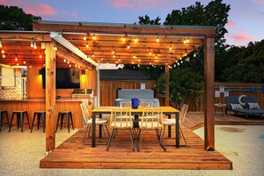 An outdoor patio with a wooden pergola, string lights, a barbecue grill, and a dining table with chairs. - 5BR Near At&t Stadium Pool, Golf Sim, EV (North Richland Hills)