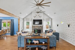 A spacious living room with a fireplace, a ceiling fan, and a large TV. - 5BR Near At&t Stadium Pool, Golf Sim, EV (North Richland Hills)