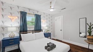 Una camera da letto con un letto, due comodini, una ventola a soffitto, una finestra con tende, una porta e uno specchio.
