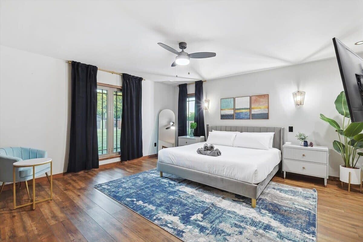 A modern bedroom with a large bed, a ceiling fan, a nightstand, a chair, and a view of the outdoors.