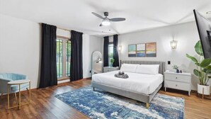 A modern bedroom with a large bed, a ceiling fan, a nightstand, a chair, and a view of the outdoors. - Russwood Acres Estate ft Gym, Cinema, Pool Table (McKinney)