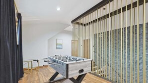 A modern room with a foosball table, wooden flooring, and a large window with vertical blinds. - Russwood Acres Estate ft Gym, Cinema, Pool Table (McKinney)