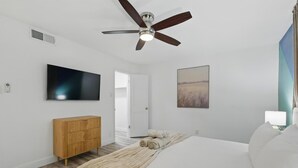Een slaapkamer met een bed, een plafondventilator, een televisie, een houten dressoir, een schilderij en een deur naar een andere kamer.