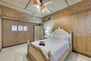 Quarto com parede de painéis de madeira, cama com roupa de cama branca, ventilador de teto e mesa de cabeceira com abajur.