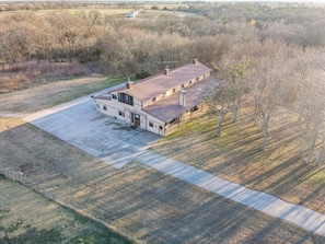 Uma casa grande e térrea, com garagem, cercada por campos abertos e algumas árvores.