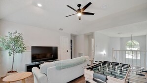 A living room with a sofa, a television, a foosball table, a ceiling fan, and a potted plant. - Tropical Pool Oasis - 5 BR, Foosbal, Cinema (Flower Mound)