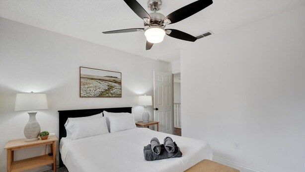 A hotel room with a bed, two bedside tables with lamps, a ceiling fan, a painting on the wall, and a bench. - Tropical Pool Oasis - 5 BR, Foosbal, Cinema (Flower Mound)