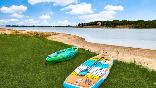 Eine grüne Wiese mit zwei Kajaks und einem Stand-Up-Paddleboard an einem ruhigen See unter blauem Himmel.