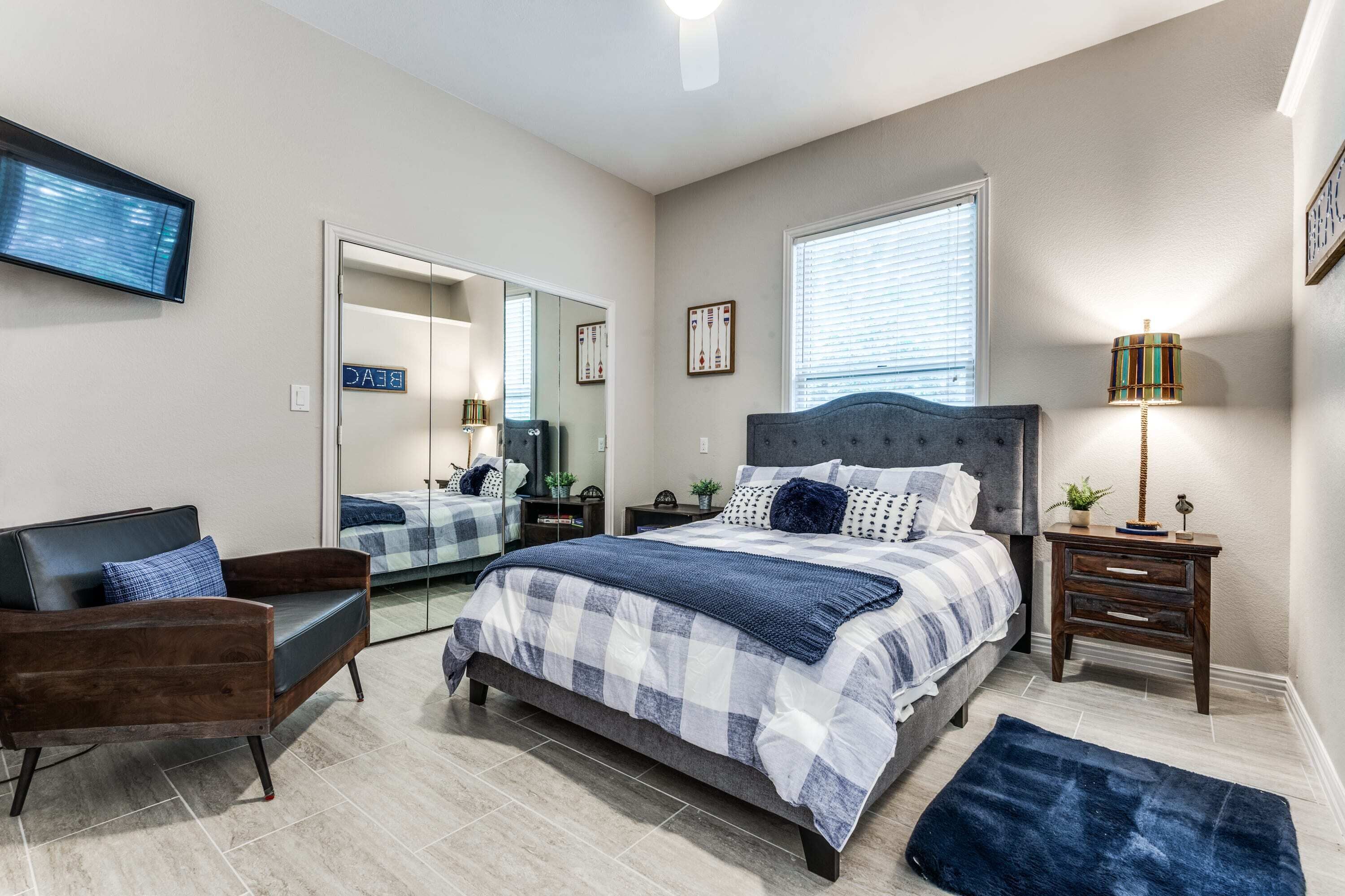 A bedroom with a bed, a chair, a nightstand, a lamp, a mirror, and a television.