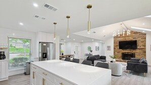 A modern kitchen with white cabinets and gold handles, a dining area with a wooden table, and a living room with a fireplace and a flat-screen TV. - 3-bd Backyard Oasis w Pool, Patio, Firepit (Irving)