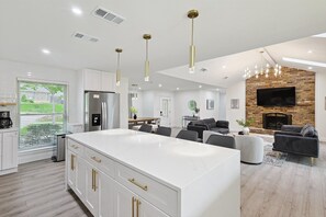 Eine moderne Küche mit weißen Schränken und goldenen Griffen, ein Essbereich mit einem Holztisch sowie ein Wohnzimmer mit Kamin und Flachbildfernseher.