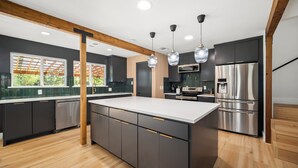 A modern kitchen with dark cabinetry, a large island, stainless steel appliances, pendant lighting, and a wooden ceiling beam. - Tranquil Country Escape, 4 Bedrooms, Pond, Firepit (Greenville)