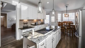 A modern kitchen with white cabinets, a granite countertop, and stainless steel appliances. There is a dining area with wooden chairs and a table. - Dream Vacay in Central Plano - Pool & Grill (Plano)