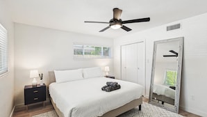 A bedroom with a bed, nightstands, a ceiling fan, a large mirror, and a window with blinds. - Bright Modern Home 3br, 2ba, Fenced backyard (Irving)