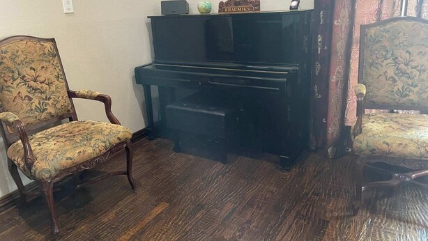 Ett rum med ett piano, två stoppade fåtöljer, ett litet bord med en namnskylt och ett vägguttag.
