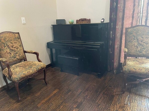 A room with a piano, two upholstered chairs, a small table with a nameplate, and a wall socket. - 5BD DFW Mansion - Pool, Cinema, Games (Plano)