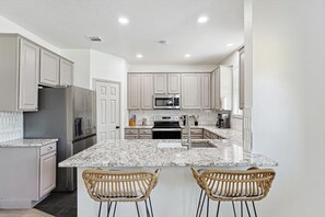 A modern kitchen with white cabinets, granite countertops, stainless steel appliances, and a central island with two wicker chairs. - Colonial Style Retreat ft Pool, Cinema, Jacuzzi (Fort Worth)
