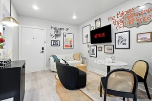 A modern living room with a white sofa, black armchair, and a round table with chairs. A flat-screen TV is mounted on the wall, and various framed artworks are displayed. - Centrally Located Cheerful 1bd/1ba Apt (Dallas)