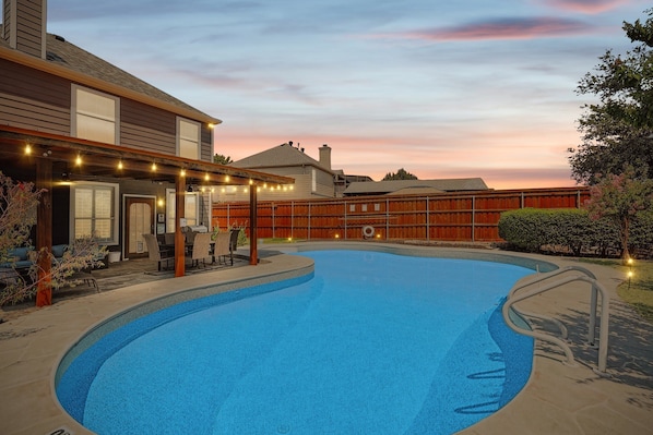 Una piscina con cubierta de madera y luces colgantes, rodeada por una cerca de madera y casas.