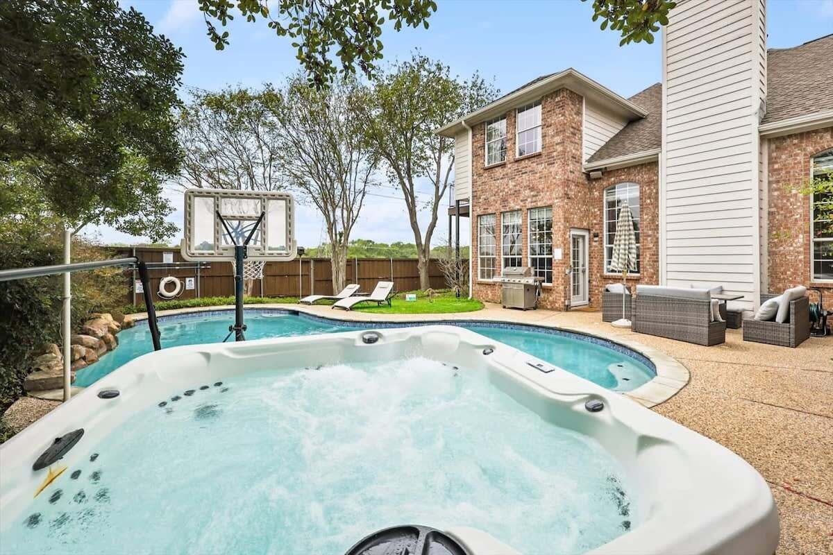 A backyard with a hot tub, a basketball hoop, and a brick house.