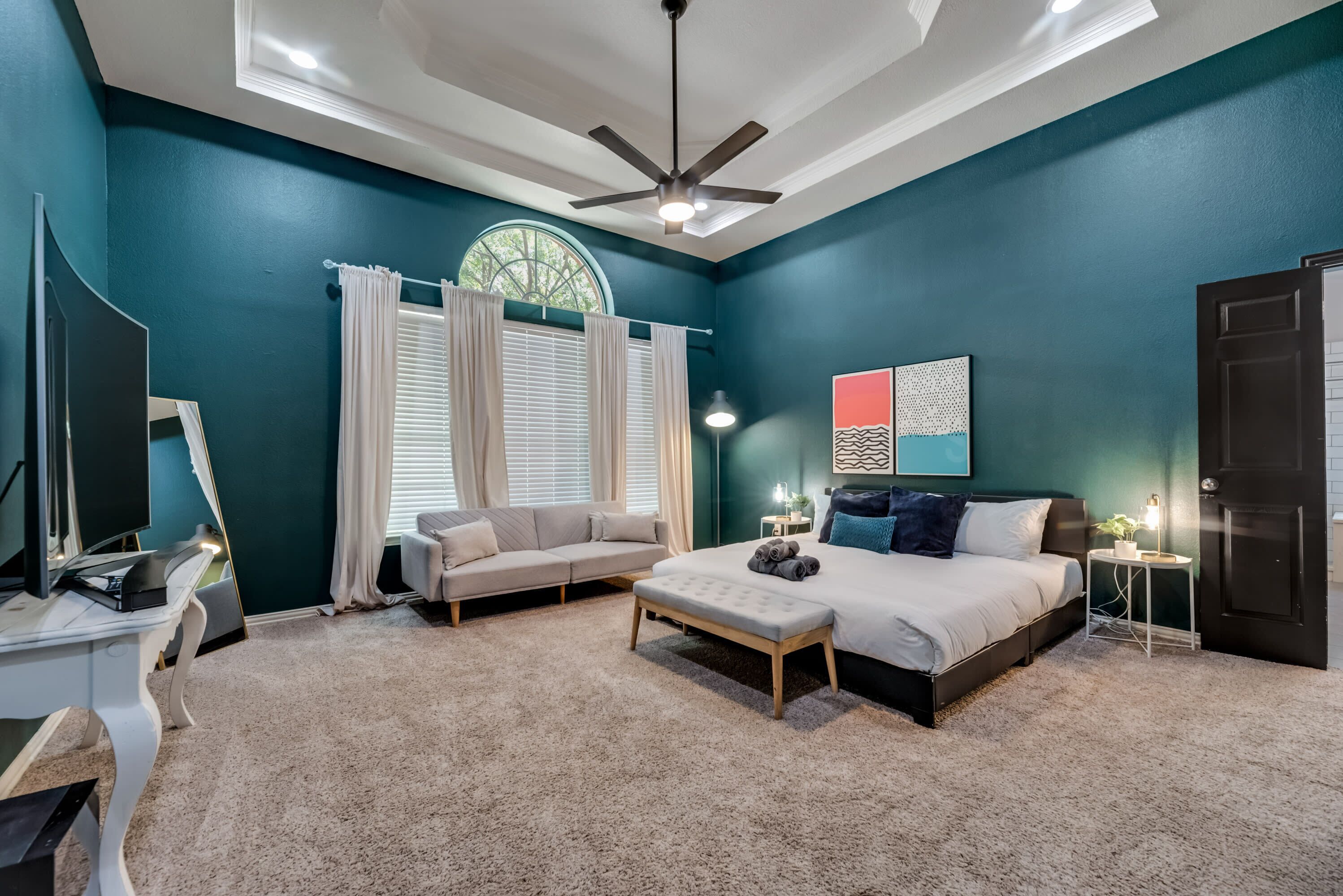 A bedroom with a bed, a desk, a TV, a ceiling fan, and a window with curtains.