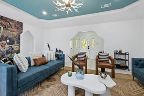 A living room with a blue sofa, two chairs, a white coffee table, and a wall with comic book art. - Country Living x Backyard Pool Oasis (Keller)