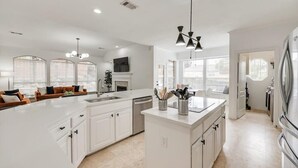 A modern kitchen with white cabinetry, a large island, and a dining area with a sofa and table. - Stunning 6br, Pool - Great for Big Groups (Rowlett)