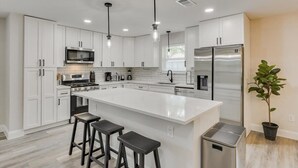 A modern kitchen with white cabinetry, stainless steel appliances, a central island with stools, pendant lighting, and a potted plant. - Gorgeous Sunny Home for Big Groups w/ Private Pool (Garland)
