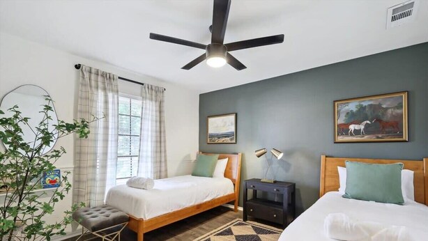 Quarto com duas camas, um ventilador de teto, mesa de cabeceira, uma planta e janela com cortinas.