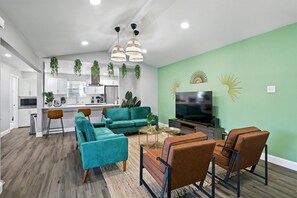 A modern living room with teal sofas, brown chairs, a TV, and hanging plants. - Best of Desoto, 5br, 2BA With Pool, Hot Tub, Patio (DeSoto)