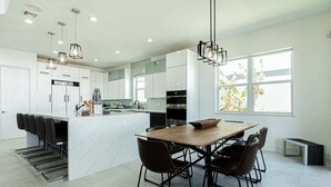 A modern kitchen with white cabinetry, a central island, and a dining area with a wooden table and chairs. - Breathtaking 6BD Riverfront Mansion w Pool (Cape Coral)