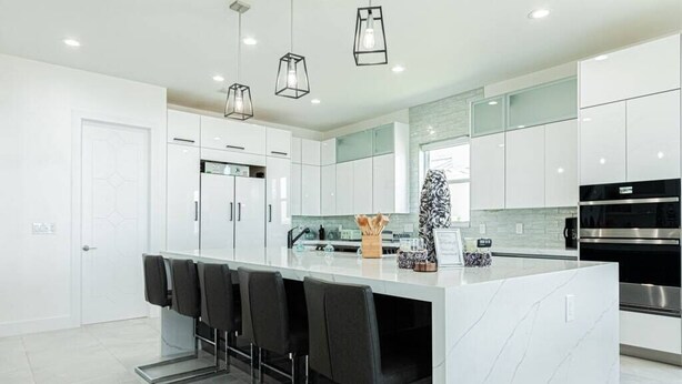 A modern kitchen with white cabinets, a marble island, black bar stools, and stainless steel appliances. - Breathtaking 6BD Riverfront Mansion w Pool (Cape Coral)