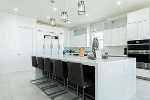 A modern kitchen with white cabinets, a marble island, black bar stools, and stainless steel appliances. - Breathtaking 6BD Riverfront Mansion w Pool (Cape Coral)