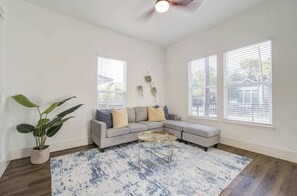 Ein Wohnzimmer mit einer Eckcouch, einem runden Couchtisch, einem Blumentopf und großen Fenstern mit Jalousien.