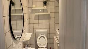 Ett badrum med vita kakelväggar, en vit toalett, ett vitt handfat, en spegel och ett rött rengöringsredskap.