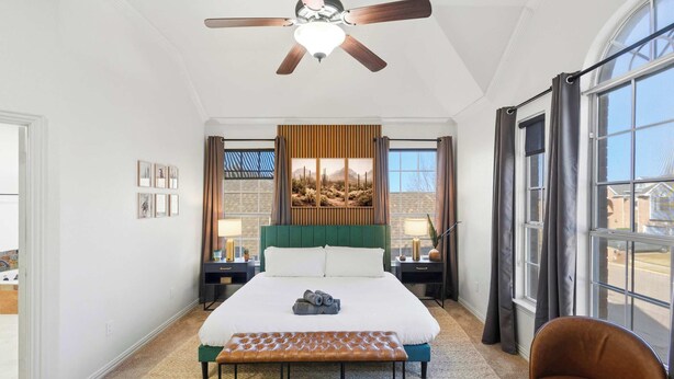 A hotel room with a large bed, a ceiling fan, a window with curtains, a bench, and a chair. - 5-bd Vacation Retreat w Pool & Ping Pong! (Fort Worth)
