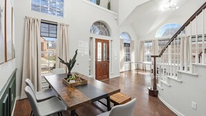 A spacious dining area with a wooden table and chairs, a staircase with white railings, and large windows with curtains. - 5-bd Vacation Retreat w Pool & Ping Pong! (Fort Worth)