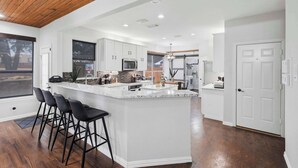 A modern kitchen with white cabinetry, a central island with bar stools, and a wooden ceiling. - 5-bd Vacation Retreat w Pool & Ping Pong! (Fort Worth)