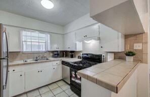 Eine moderne Küche mit weißen Schränken, einem Edelstahl-Kühlschrank, einem Gasherd und einem Fenster mit Jalousien.