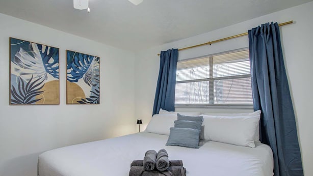 A neatly made bed with a rolled towel, two wall art pieces, and blue curtains. - Heart of Houston - 3-bd Apartment (Houston)