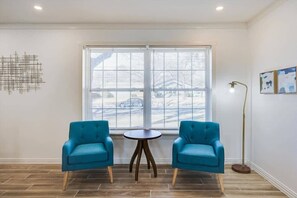 Ein Zimmer mit zwei blauen Sesseln, einem kleinen runden Tisch, einer Stehlampe und einem Fenster mit Jalousien.