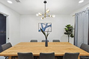 Un salón de conferencias con una mesa de madera, sillas grises, una lámpara de araña moderna, un cuadro abstracto enmarcado y una planta en maceta.
