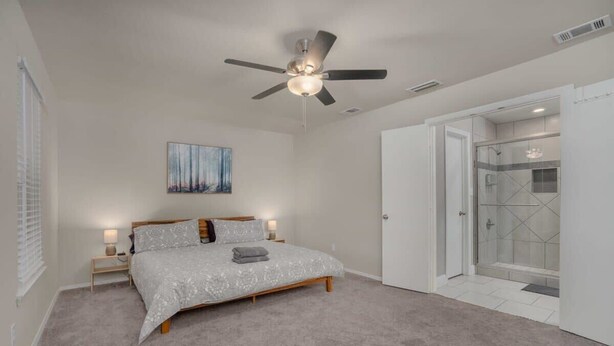 Un dormitorio con cama, ventilador de techo, un cuadro, mesita de noche y un vestidor que da acceso a la zona de la ducha.