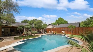 A backyard with a swimming pool, surrounded by a wooden fence, a patio with outdoor furniture, and various trees and shrubs. - Highland Village - 4BR w Backyard Pool Oasis (Lewisville)